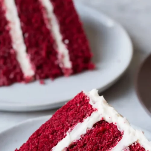 A slice of red velvet cake generously topped with cream cheese frosting, showing its vibrant red layers.